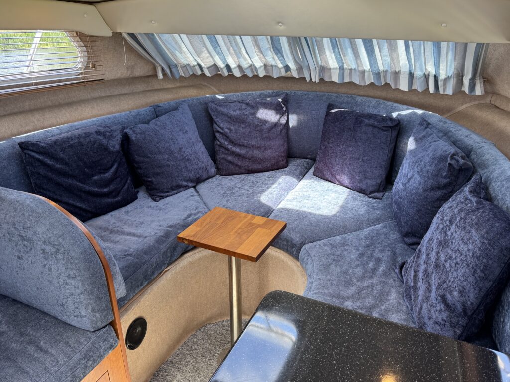 A cozy seating area with a circular arrangement of dark blue cushions against a beige wall. A small wooden table stands in the center, while light streams through striped curtains above.