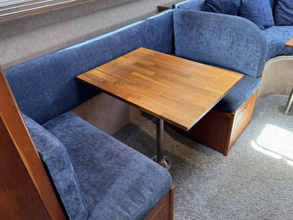 A cozy seating area featuring a wooden table supported by a metal pedestal, surrounded by upholstered blue couches with multiple cushions. The background includes a light-colored wall with window blinds and soft carpeting.