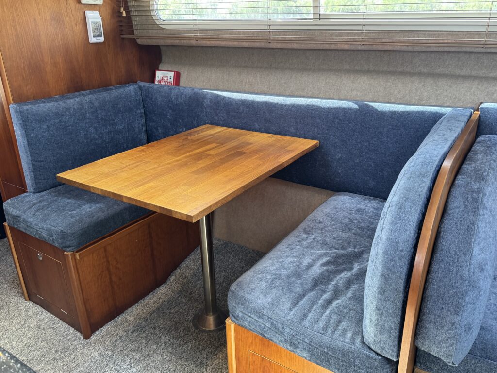 A small dining area featuring a wooden table with a square top supported by a central pedestal. The space includes a U-shaped bench covered in blue fabric, with light streaming through a window in the background. Wooden cabinetry is also visible.