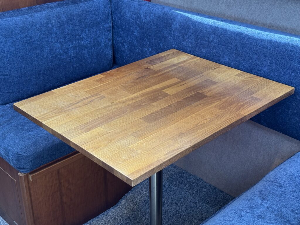 A wooden square table with a smooth finish is positioned between blue cushioned seats in a corner seating area. The table features visible wood grain and is supported by a metal leg.