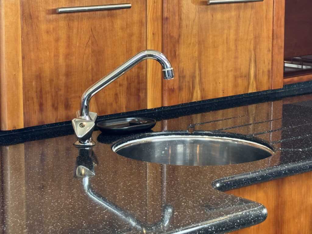 A polished black countertop features a stainless steel sink and a curved faucet. Behind the countertop, there are wooden cabinets with sleek handles visible.