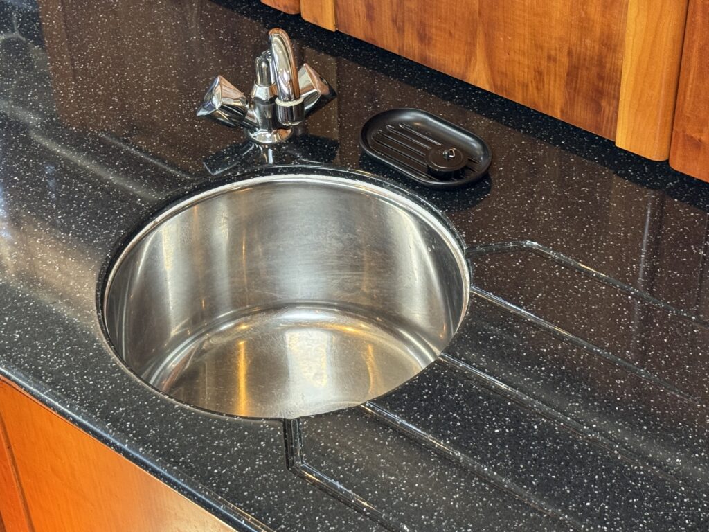 A stainless steel sink with a circular basin is set in a black speckled countertop. Above the sink, a chrome faucet is positioned, and beside it, a black dish for soap sits on the countertop.