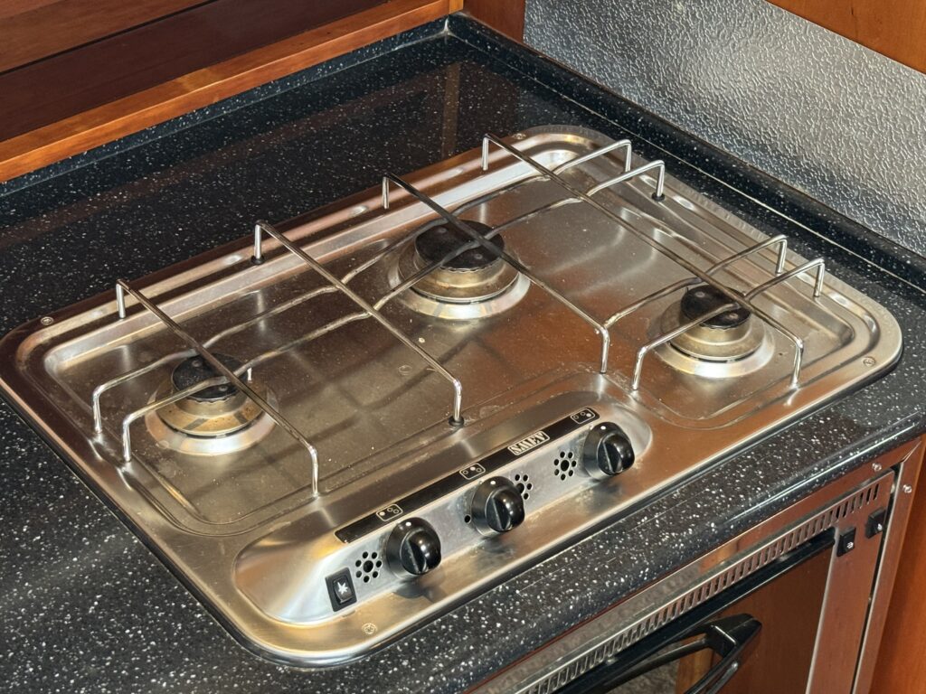 A stainless steel gas stovetop with three burners and control knobs located at the front. The surface is set into a dark speckled countertop, with metal grates over the burners, showcasing a modern kitchen design.