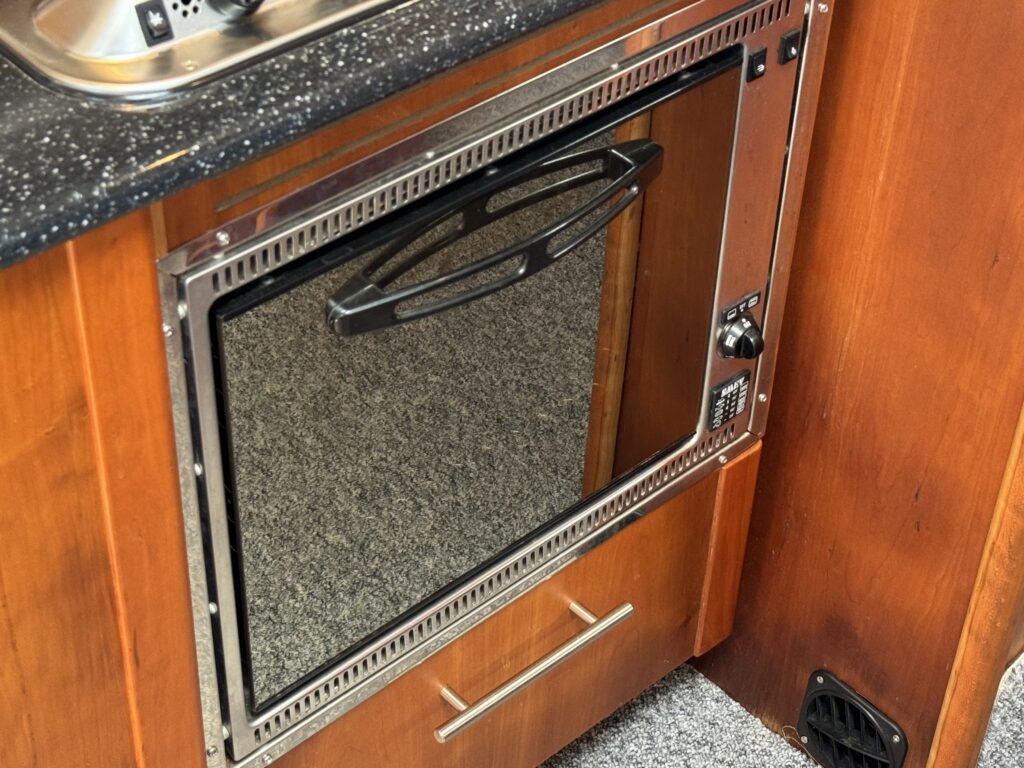 A built-in kitchen oven with a stainless steel and glass door, set into a wooden cabinet. The oven features a handle for opening and has a control panel on the right side, with a grey carpeted floor visible below.