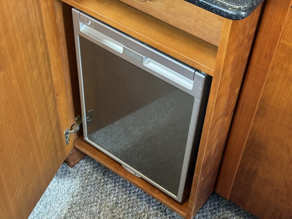 A silver compact refrigerator is built into a wooden cabinet with a visible grain finish. The refrigerator door is partially closed, revealing a reflective surface. The countertop above is black with a speckled pattern.