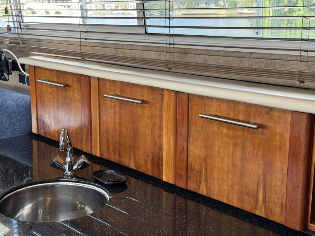 A kitchen countertop featuring three wooden cabinet doors with metallic handles beneath a sleek solid surface. A silver sink is visible to the left, and natural light filters through a window with horizontal blinds, reflecting off a water surface outside.