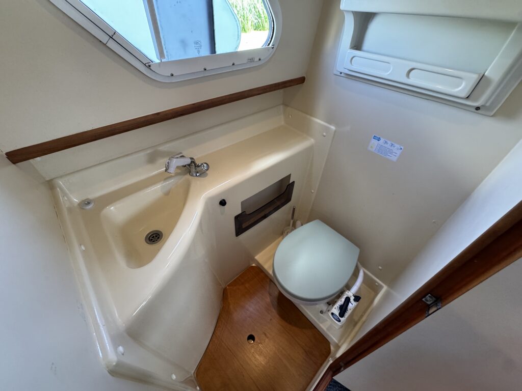 A small boat bathroom featuring a compact toilet and a sink with a single faucet. The walls are white, and there is a window providing natural light. Wooden accents are visible near the sink and along the floor.