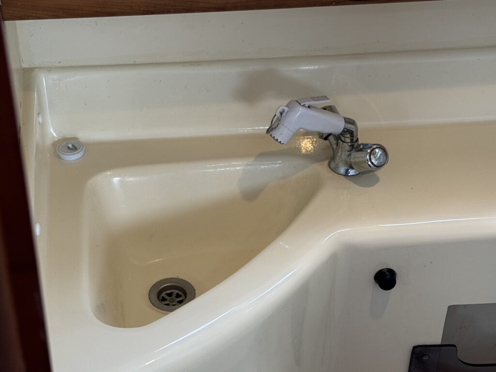 A close-up of a white sink featuring a chrome faucet with a white lever handle. The sink has a rounded corner, and a drain is visible at the bottom left. The surface is smooth and lightly textured, with a surrounding wooden edge.