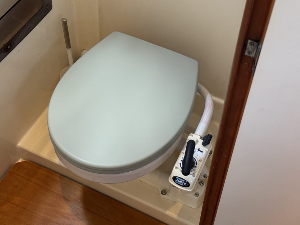 A compact marine toilet with a light blue seat is positioned in a small, enclosed space. A control pump is attached to the side, and the floor is made of wood, enhancing the compact design of the area.