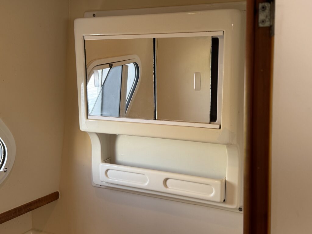 A white, mounted bathroom mirror with a sliding panel below it. The mirror has a square shape, reflecting light and showing part of a window in the background. The surrounding wall is beige, and there is a small, wooden ledge beside the mirror.