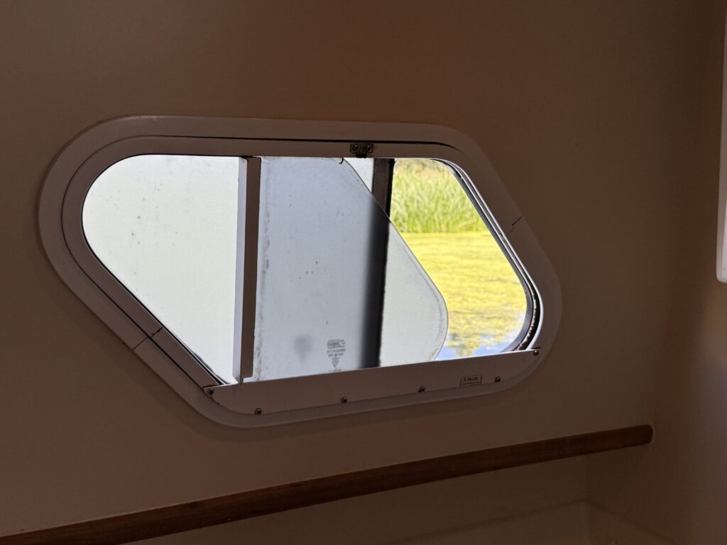 An oval-shaped window in a boat cabin is shown, partially open to reveal a view of grassy greenery outside. The interior wall is a light color, and a wooden ledge runs beneath the window.