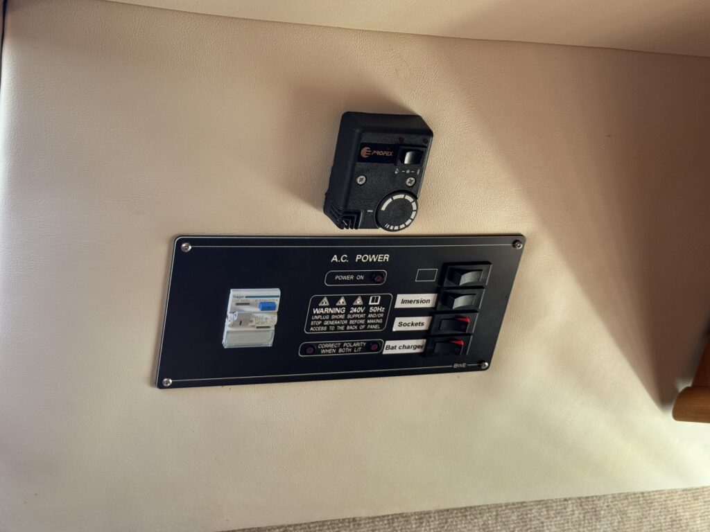 A control panel mounted on a beige wall featuring an A.C. power switch, a warning label, and various labeled switches for different functions, including sockets and charging options. A small black device with a dial is positioned above the panel.