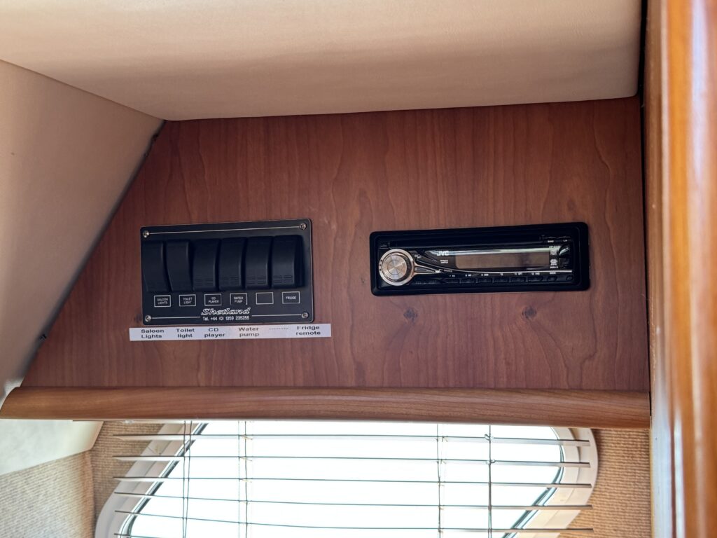 A wooden panel featuring an array of switches on the left, labeled for different functions, and a radio or audio system on the right. The background includes a section of a window with horizontal blinds, allowing some light into the area.