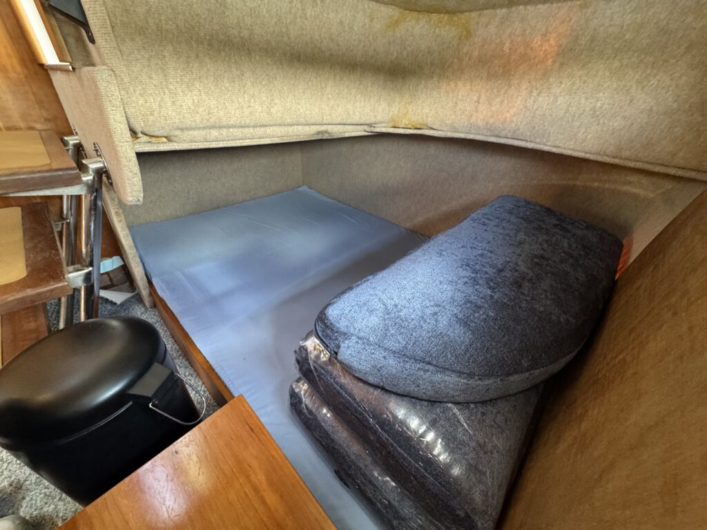 A compact sleeping area featuring a gray cushioned mattress, stacked cushions, and wooden surfaces. The background shows a padded wall with a foldable upper section, creating a cozy, enclosed space.