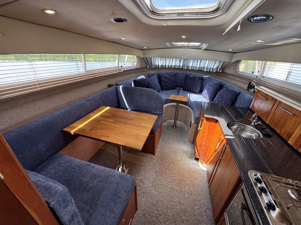 An interior of a boat featuring a comfortable seating area with a curved blue couch and several cushions. A wooden table is positioned near the couch, and the kitchenette is visible with a sink and a stove, all under natural light from windows.