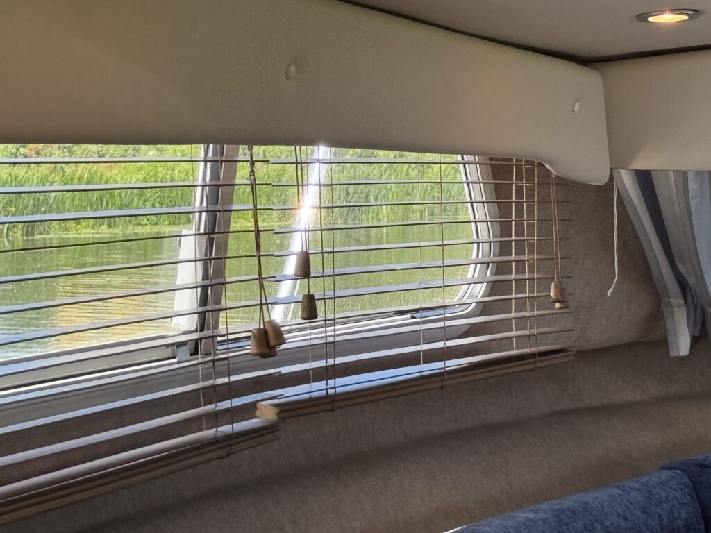 Interior view of a boat with vertical blinds covering a window. The blinds allow a view of green foliage reflected in calm water outside. Light enters the space, and a soft beige and blue color palette is visible in the decor.