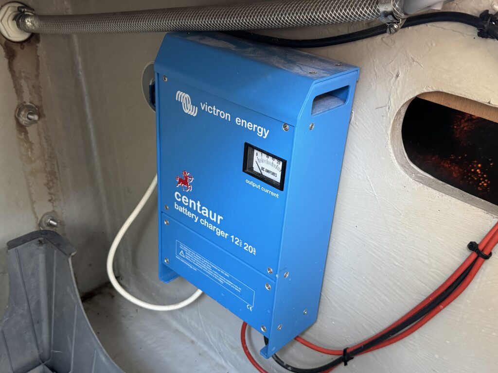 A blue Victron Energy Centaur battery charger mounted on a wall. The charger features a digital display showing output status, with cables attached in a neat arrangement. Nearby, a gray compartment is visible, along with exposed wiring.