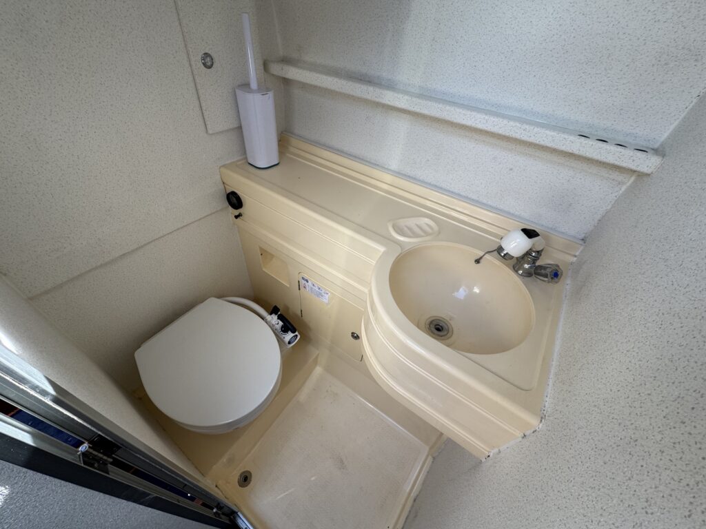 A compact bathroom area featuring a white toilet, a beige sink with a single faucet, and a soap dispenser. The walls and surfaces are light-colored and smooth, contributing to a clean, utilitarian appearance.