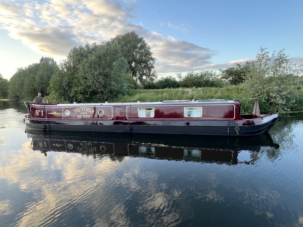A burgundy narrowboat named "Victoria" is floating on calm water, surrounded by lush greenery and trees. The boat features several windows, and the sky above is partly cloudy, reflecting in the water.