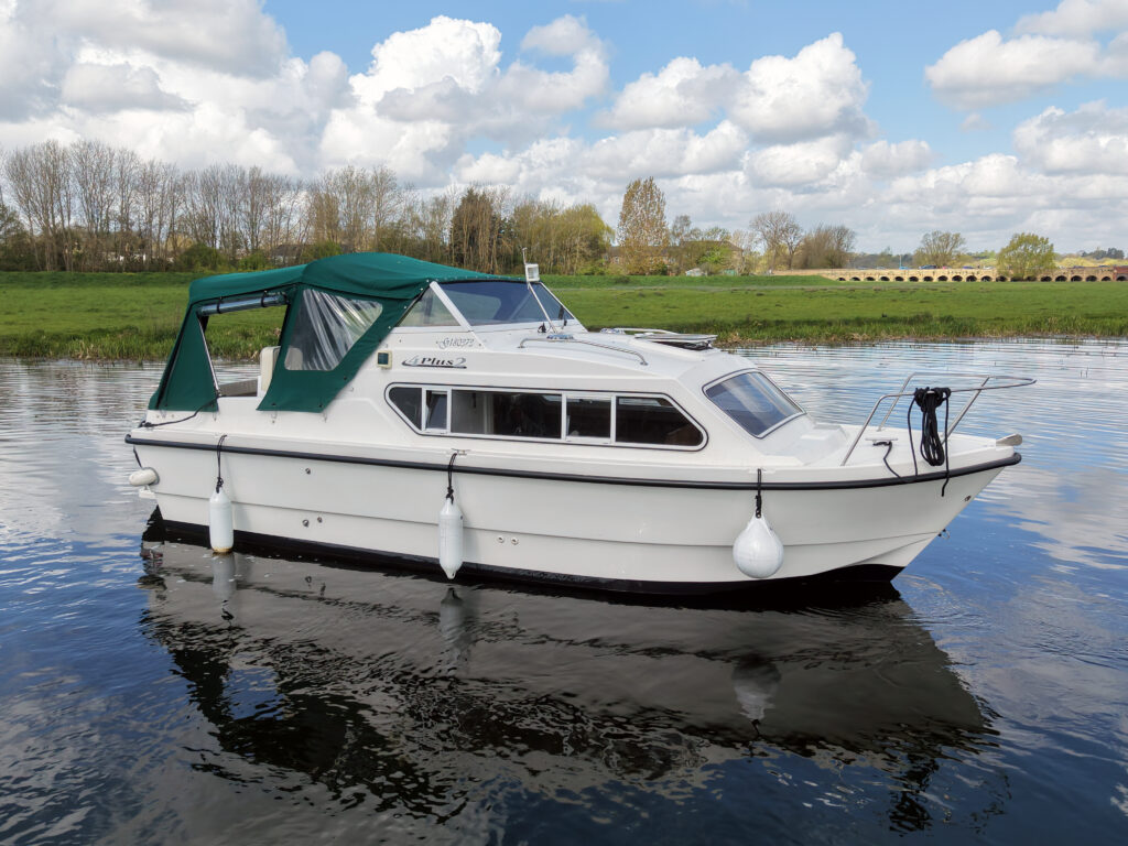 A white motorboat with a green canopy is moored on calm water. The boat features multiple windows and fenders, with trees and clouds visible in the background.