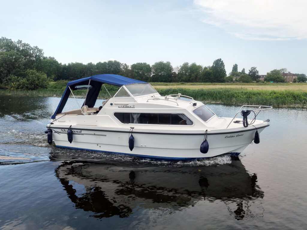 A white motorboat with a blue canopy is cruising on calm waters. The background features lush greenery and trees along the shore, with a clear sky overhead.