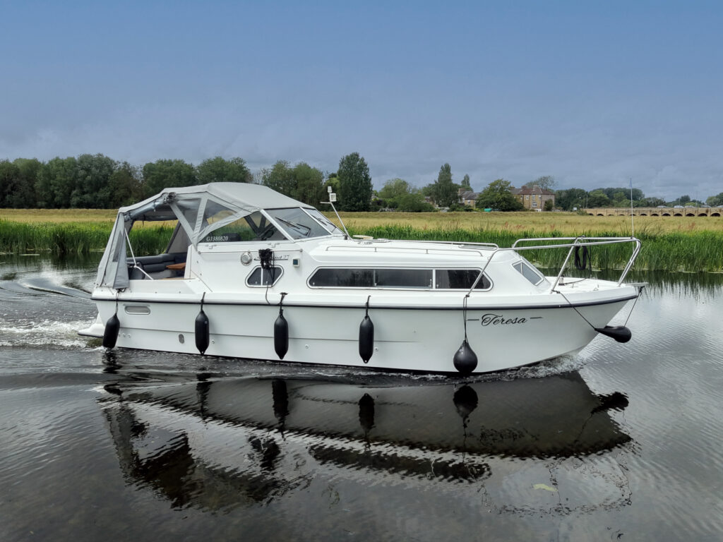 A white cabin cruiser boat named 'Teresa' sails on calm waters. The boat features a canopy and several fenders along the sides, with lush green trees and a field visible in the background under a cloudy sky.