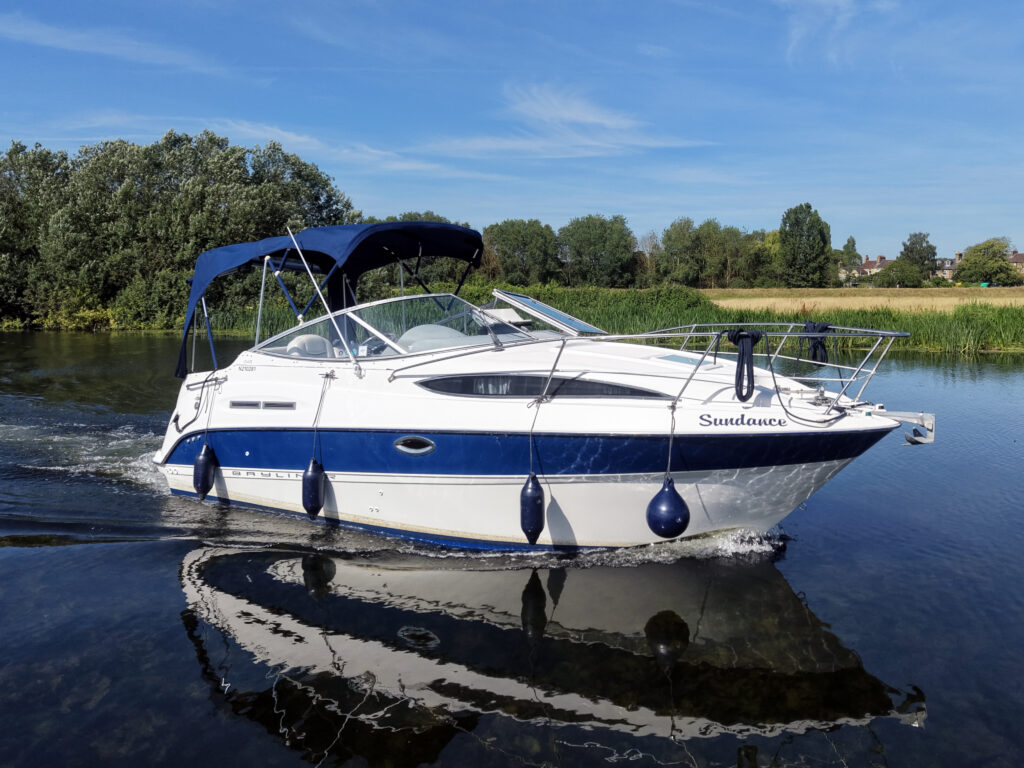 A white and blue motorboat named "Sundancer" is cruising on a calm waterway. The boat features a canopy and several fenders hanging over the side, with lush greenery visible along the shore in the background.