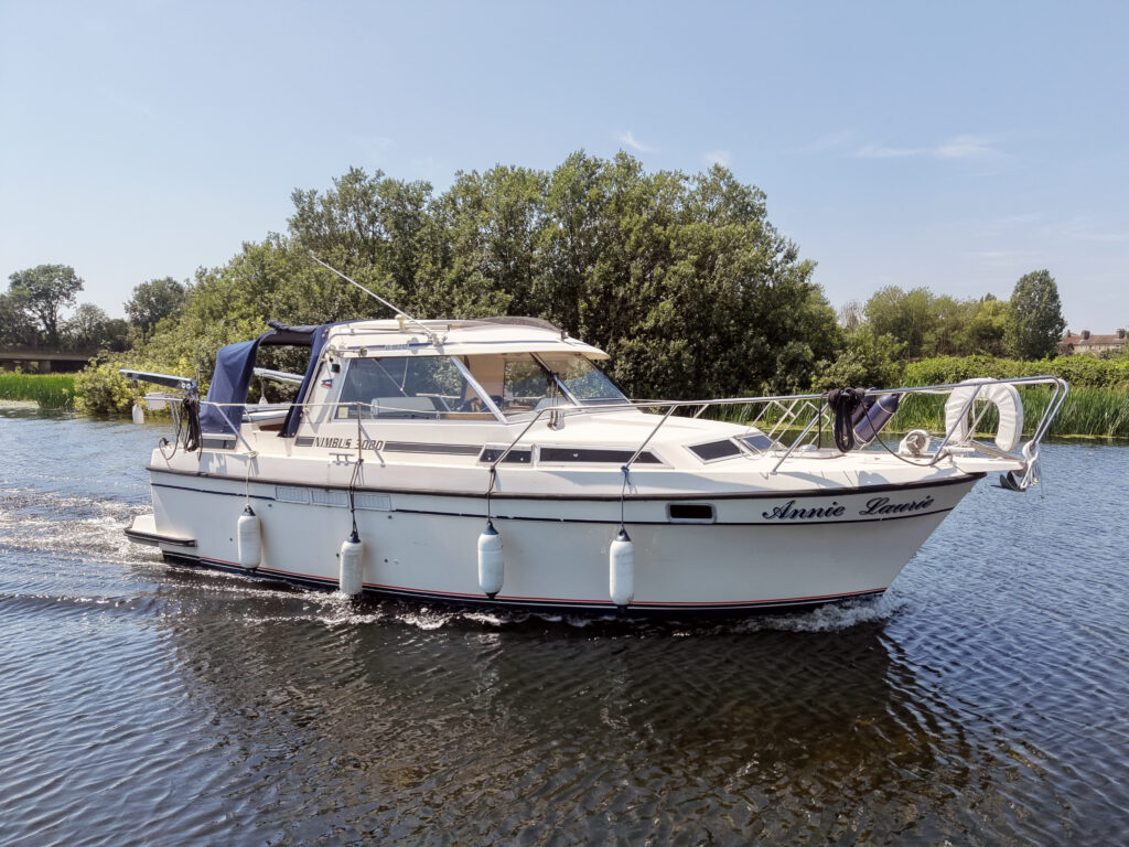 A white motorboat named "Annie Louise" is navigating through calm waters surrounded by greenery. The boat features a covered cockpit area and is equipped with several fenders hanging from the side.