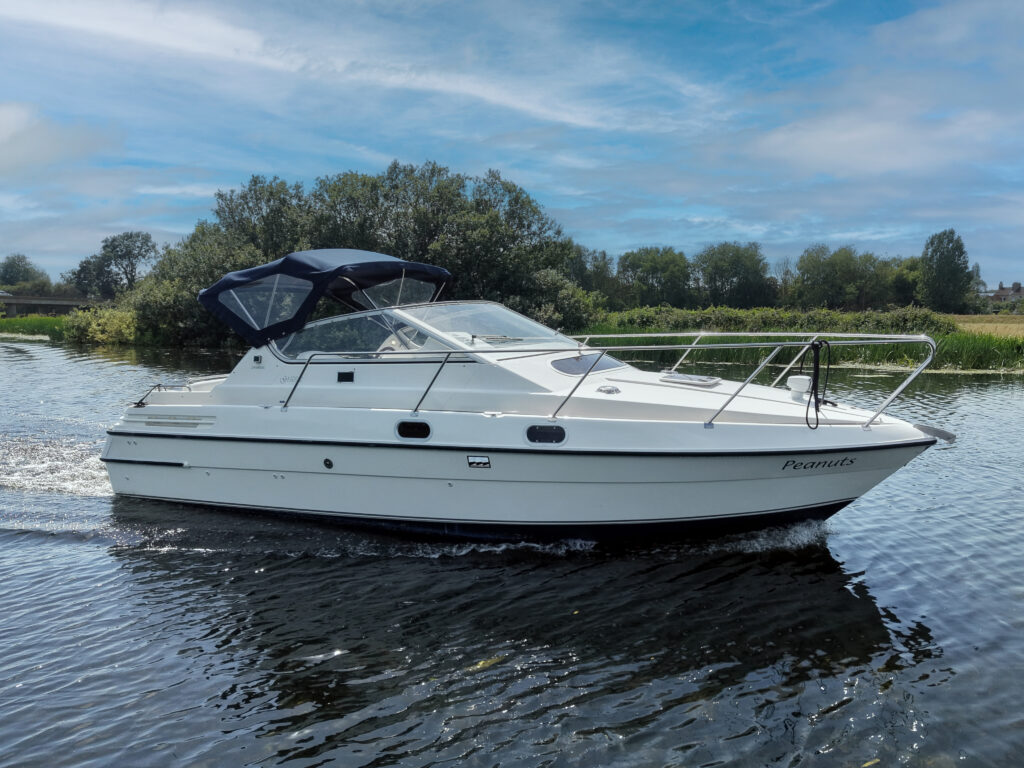 A white cabin cruiser boat with a blue canopy is navigating through a calm river. Lush greenery borders the water, and a clear blue sky is visible above.