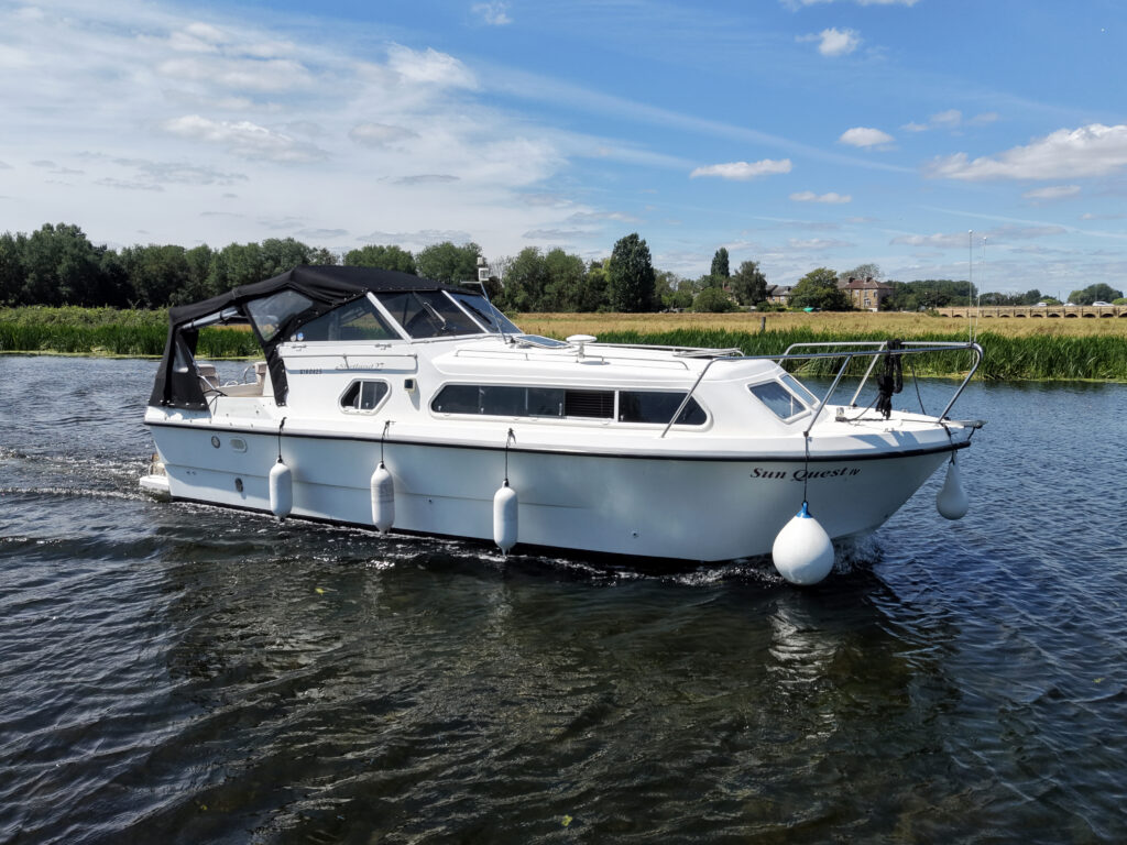 A white motorboat with a black canopy moves through calm water. It is anchored by two white buoys and surrounded by green grass and trees in the background under a clear blue sky.