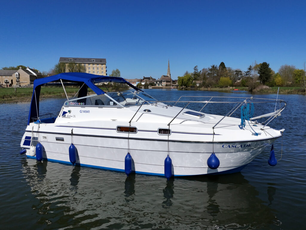A white motorboat with blue accents is anchored in calm water. The boat features a blue canopy and several blue fenders. In the background, buildings and trees are visible, under a clear blue sky.