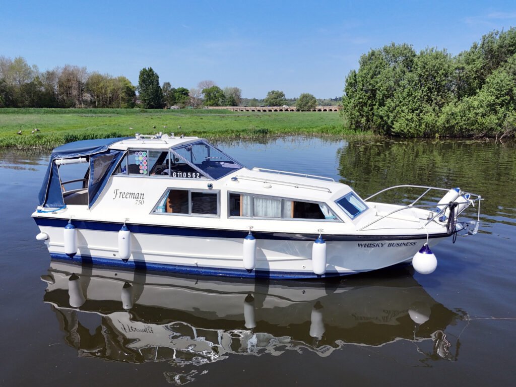A white motorboat named "Freeman" is moored on calm water, surrounded by greenery and trees. The boat features a blue canopy and is equipped with a railing and fender on the bow. The landscape includes a bright blue sky.