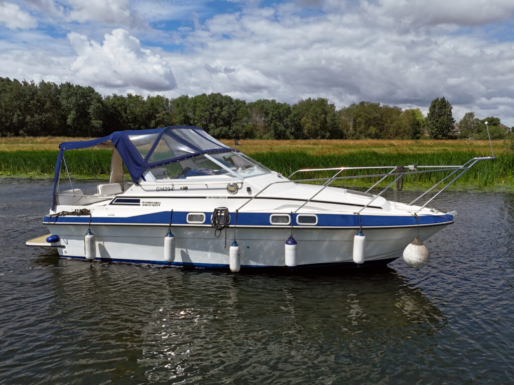 A white and blue motorboat anchored on calm water, featuring a clear windshield and a blue canopy. Surrounding greenery and a cloudy sky are visible in the background.