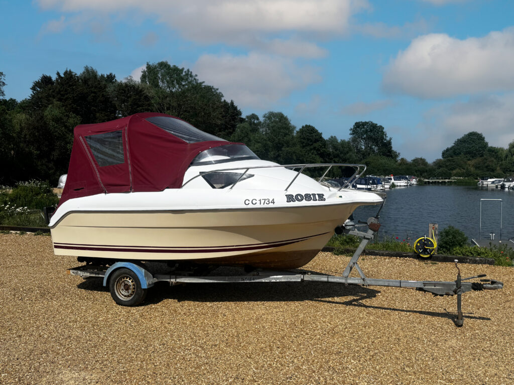 A white and beige Quicksilver 510 boat with a maroon canopy is secured on a trailer next to a calm body of water. The scene includes trees in the background and a clear blue sky with a few clouds.