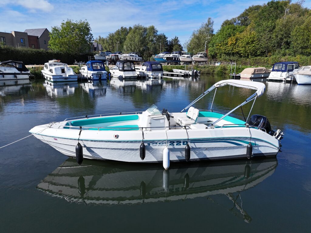 A white motorboat with a turquoise interior is anchored on calm water in a marina. Several other boats are visible in the background, along with green trees and a clear sky.