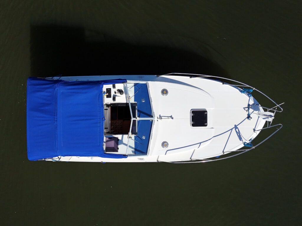 An aerial view of a white motorboat with a blue canopy. The boat has a small open deck with visible seating, a steering wheel, and a cabin area. The water surrounding the boat is calm and dark.
