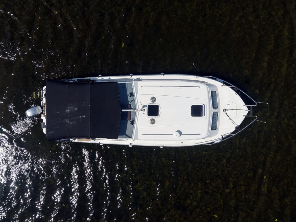 An aerial view of a white motorboat with a black canopy on the surface of a dark water body. The boat features a cabin with square windows and is surrounded by reflections of sunlight on the water.