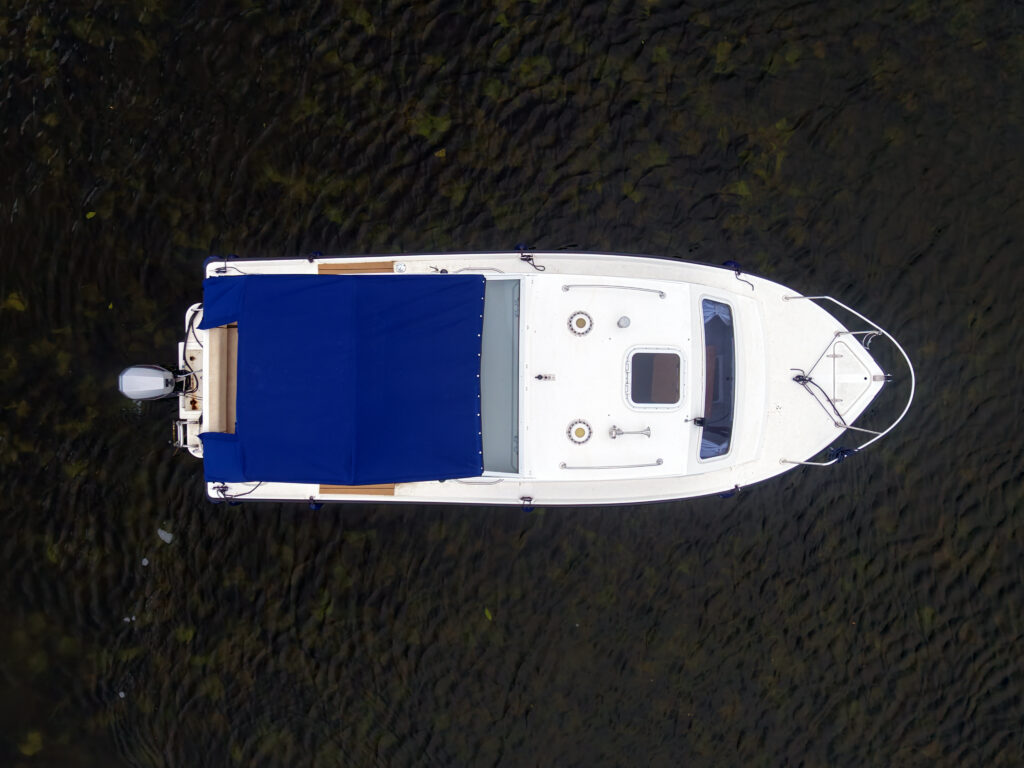 An aerial view of a white boat with a blue canopy on a body of water. The boat features a small cabin, an outboard motor, and various deck fittings visible on the surface.