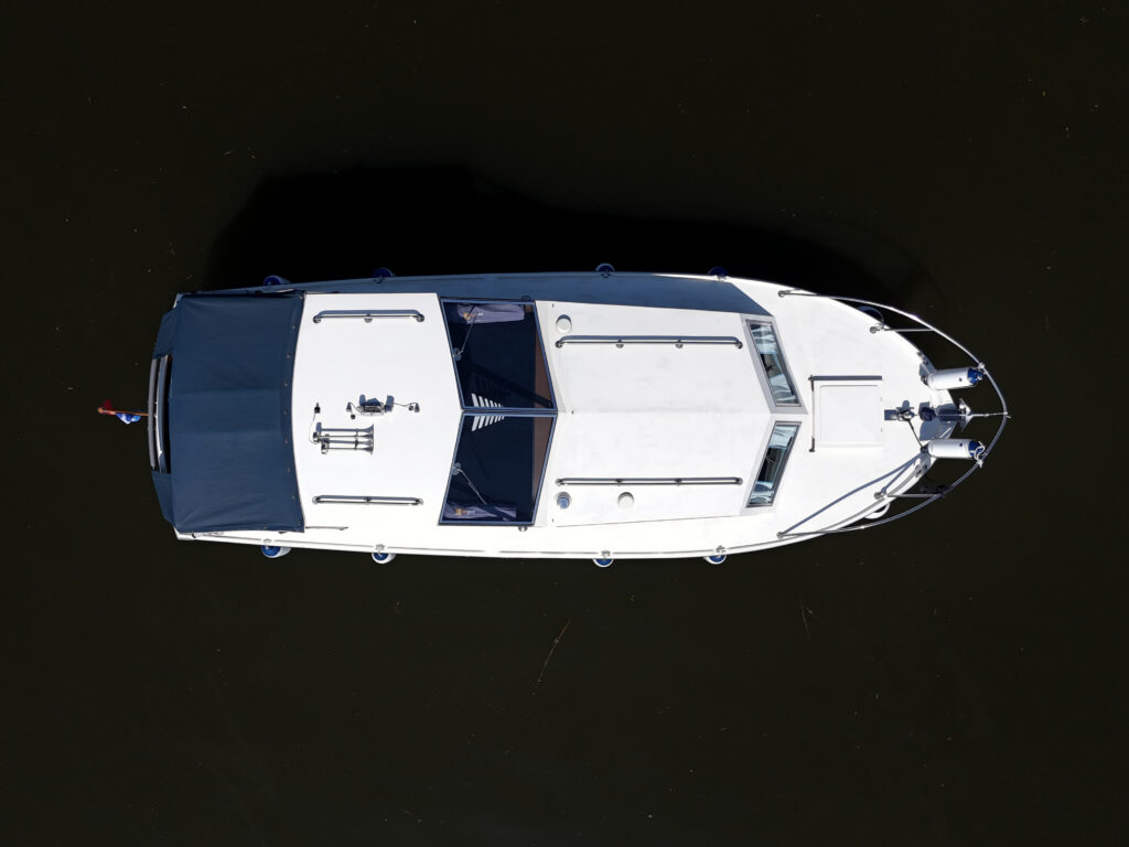 An overhead view of a white motorboat with a dark blue cover on its bow, moored on still water. The boat features multiple windows and an open deck space.