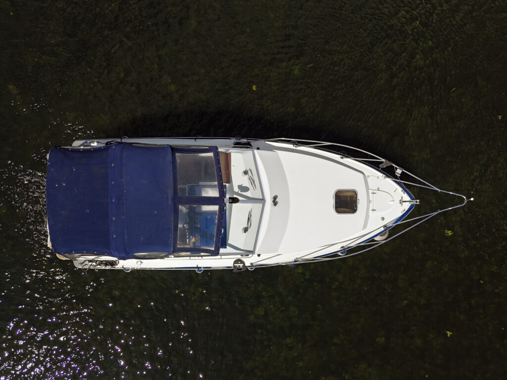 An aerial view of a white motorboat with a blue canopy docked on calm water. The boat features a cabin with windows and is surrounded by the reflection of natural surroundings in the water.