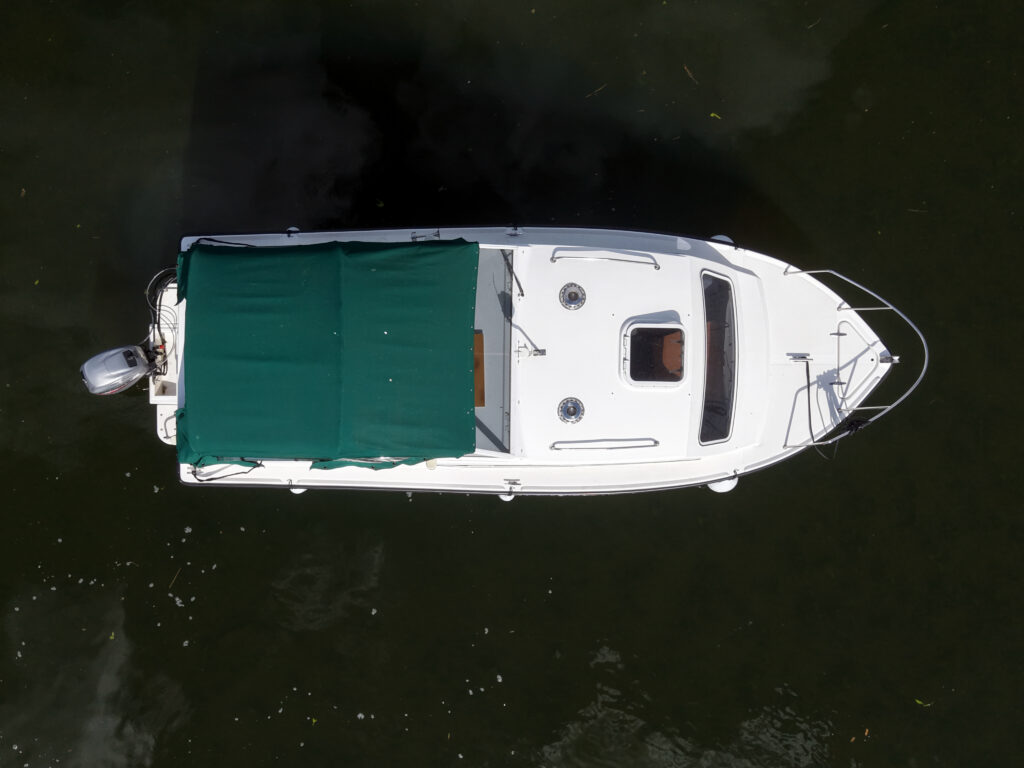 An aerial view of a white boat with a green canopy covering the cabin area. The boat is positioned on calm water, with its propeller visible at the rear and several circular hatches on the deck.
