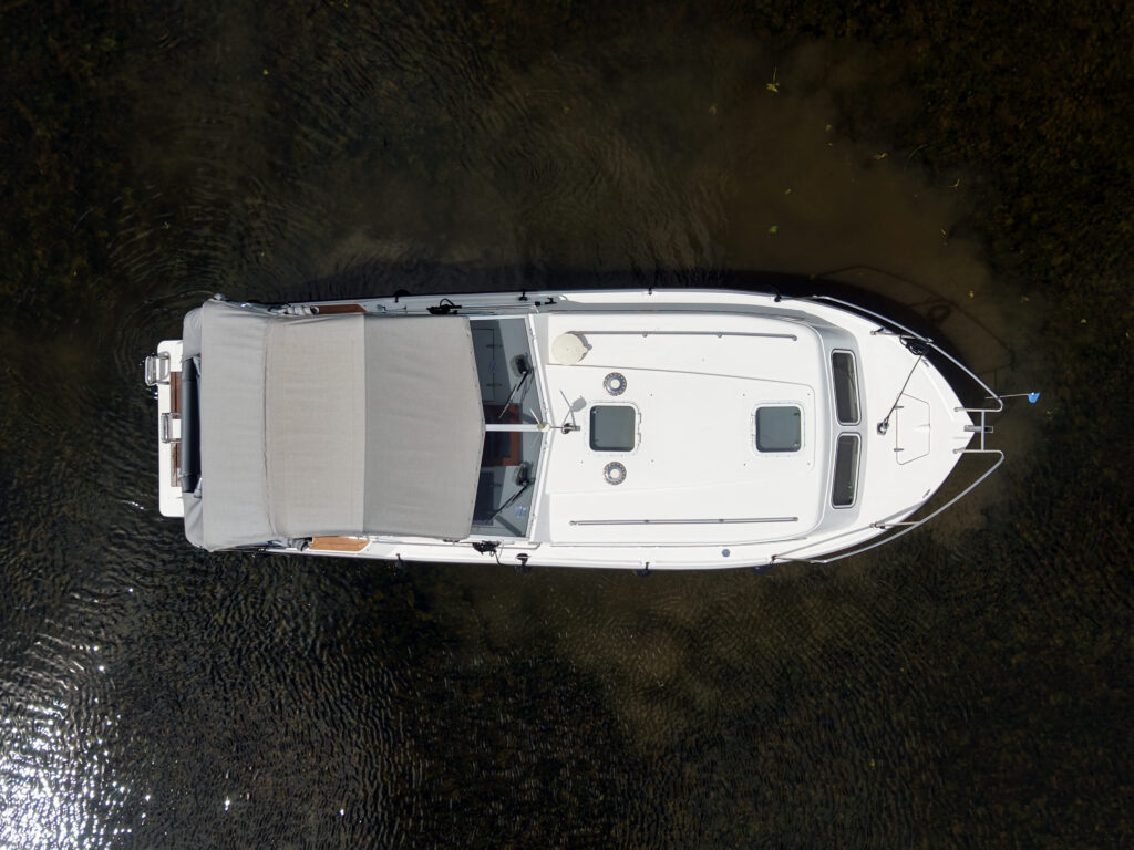 An aerial view of a white motorboat with a covered area on the deck, anchored in calm water. The boat features large windows and a blue flag at the bow, with sunlight reflecting on the water's surface.