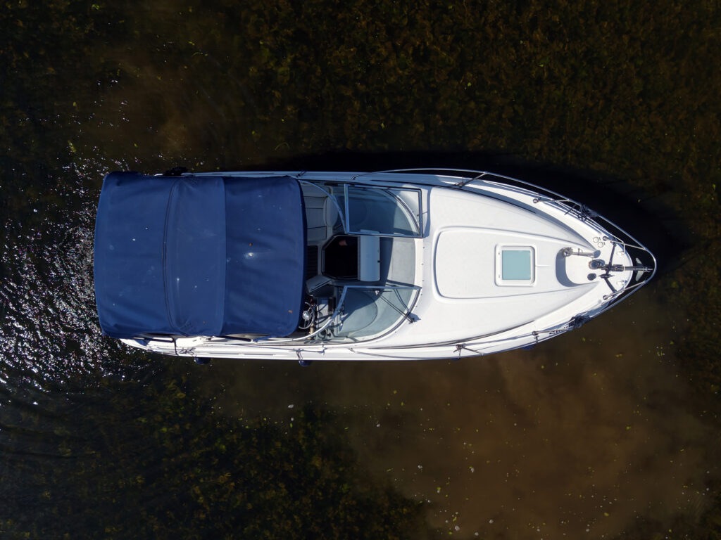 An aerial view of a white motorboat with a blue canopy on the water. The boat has a clear windshield and a small cabin area; the water below features some visibility of the riverbed.