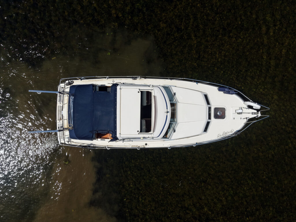 An aerial view of a white motorboat with a blue cover, anchored on shallow water. The boat features a streamlined design and has seats visible on deck. The surrounding water has a dark green appearance, providing contrast to the boat.