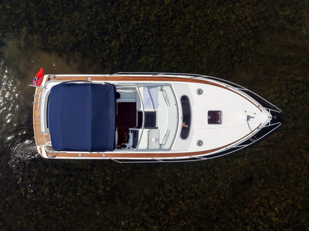 An aerial view of a white motorboat with a blue canopy and wooden accents, anchored on clear water. The boat features a small flag at the rear and is surrounded by a natural landscape reflected in the water.