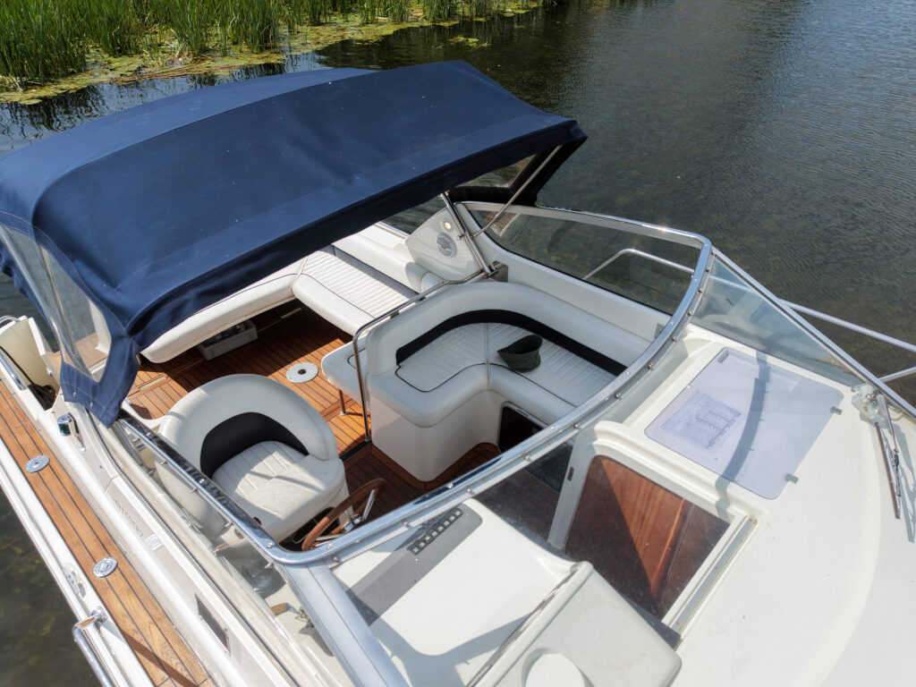 An overhead view of a white boat with a navy blue canopy. The interior features light-colored seating arranged around a central table, and is set atop a wooden deck. Surrounding the boat are green plants and calm water.