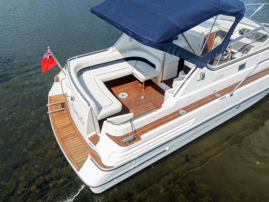 An aerial view of a white motorboat anchored in shallow water. The boat features a dark blue canopy, wooden deck accents, and a red flag. Cushioned seating is positioned at the rear, complementing the boat's clean design.