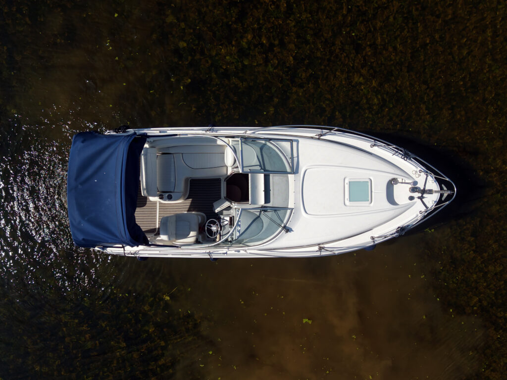 An overhead view of a white motorboat with a blue canopy, docked in calm water. The boat features cushioned seating and a steering wheel at the front. The surrounding water has a dark green hue from the vegetation below.