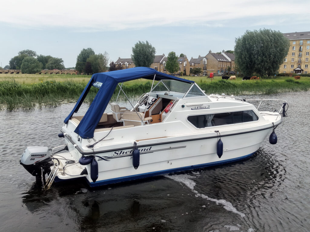 A white motorboat with a blue canopy is moored on calm water. The boat features a small cabin and is surrounded by lush greenery and buildings in the background.