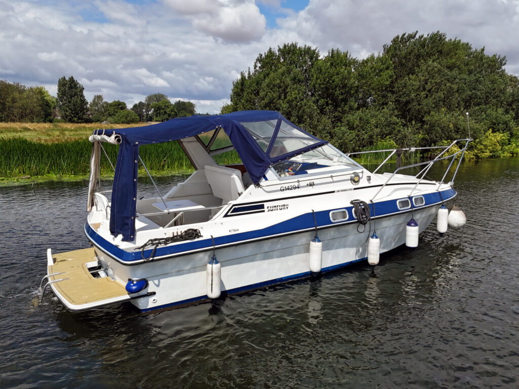 A Fairline Sunfury boat is moored on calm waters, featuring a blue and white exterior. It has a canopy above the cabin and is surrounded by greenery and trees under a partly cloudy sky.